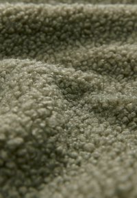 Textureret olivengrønt stof med en fuzzy, blød strikket overflade, der har et hævet løbepattern. Materialet ser tykkere og hyggeligere ud.