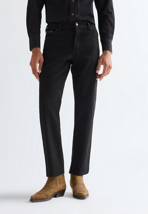 Mann trägt schwarze Jeans mit geradem Bein, schwarzes Hemd und beige Wildleder-Stiefeletten, steht vor einem schlichten weißen Hintergrund.