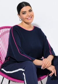 Frau mit dunklem, nach hinten gebundenem Haar, trägt ein dunkelblaues Outfit mit rosa und weißen Streifen, sitzt auf einem hellpinkfarbenen Stuhl und lächelt in die Kamera.
