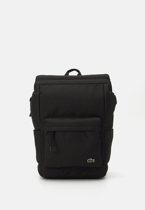 Mochila negra, hecha de un tejido duradero, cuenta con un gran compartimento principal, bolsillo frontal con cremallera, bolsillos laterales y un asa en la parte superior. Logotipo de Lacoste visible.