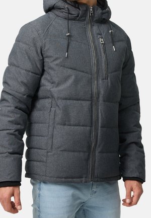 Homme portant une veste matelassée gris foncé avec capuche, fermeture éclair frontale, poche poitrine zippée, et jean bleu clair sur fond blanc.
