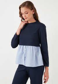 Maglietta a maniche lunghe blu navy con dettagli a peplum a righe blu e bianche, scollatura rotonda e dettagli con bottone sul davanti. Tessuto liscio.