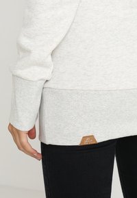 Vaaleanharmaa collegepaita, jossa on ribbireunat hihoissa ja helmassa, varustettuna nahkaisella logolapulla alaosassa. Pehmeä kangas näkyy tekstuurissa.