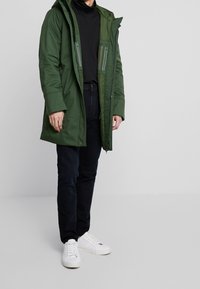 Parka verde con capucha, con bolsillos con cremallera, de textura suave y largo hasta la mitad del muslo. Llevado sobre un jersey negro de cuello alto y jeans oscuros.
