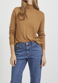 Maglione dolcevita color cammello in un tessuto morbido, abbinato a jeans in denim a vita alta di una tonalità blu media. Design semplice, aderente e maniche lunghe.