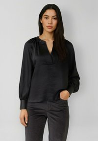 Frau mit langen dunklen Haaren, die eine schwarze Satinbluse mit Puffärmeln und dunkle Hosen trägt, steht vor einem schlichten hellen Hintergrund.