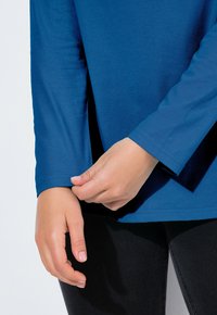 Niebieska bluzka z długim rękawem wykonana z gładkiej tkaniny, z dopasowanymi rękawami, bocznymi rozcięciami i okrągłym dekoltem. Ręka poprawia mankiet rękawa.