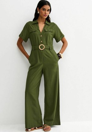 Femme portant une combinaison pantalon ample vert olive à manches courtes, avec des boutons, une ceinture à boucle ronde et des sandales, debout contre un mur blanc.