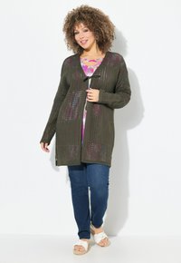 Cardigan lavorato a maglia verde oliva con trama aperta, chiusura davanti con lacci, tasche, indossato sopra una maglietta rosa a fantasia e abbinato a jeans blu e sandali.