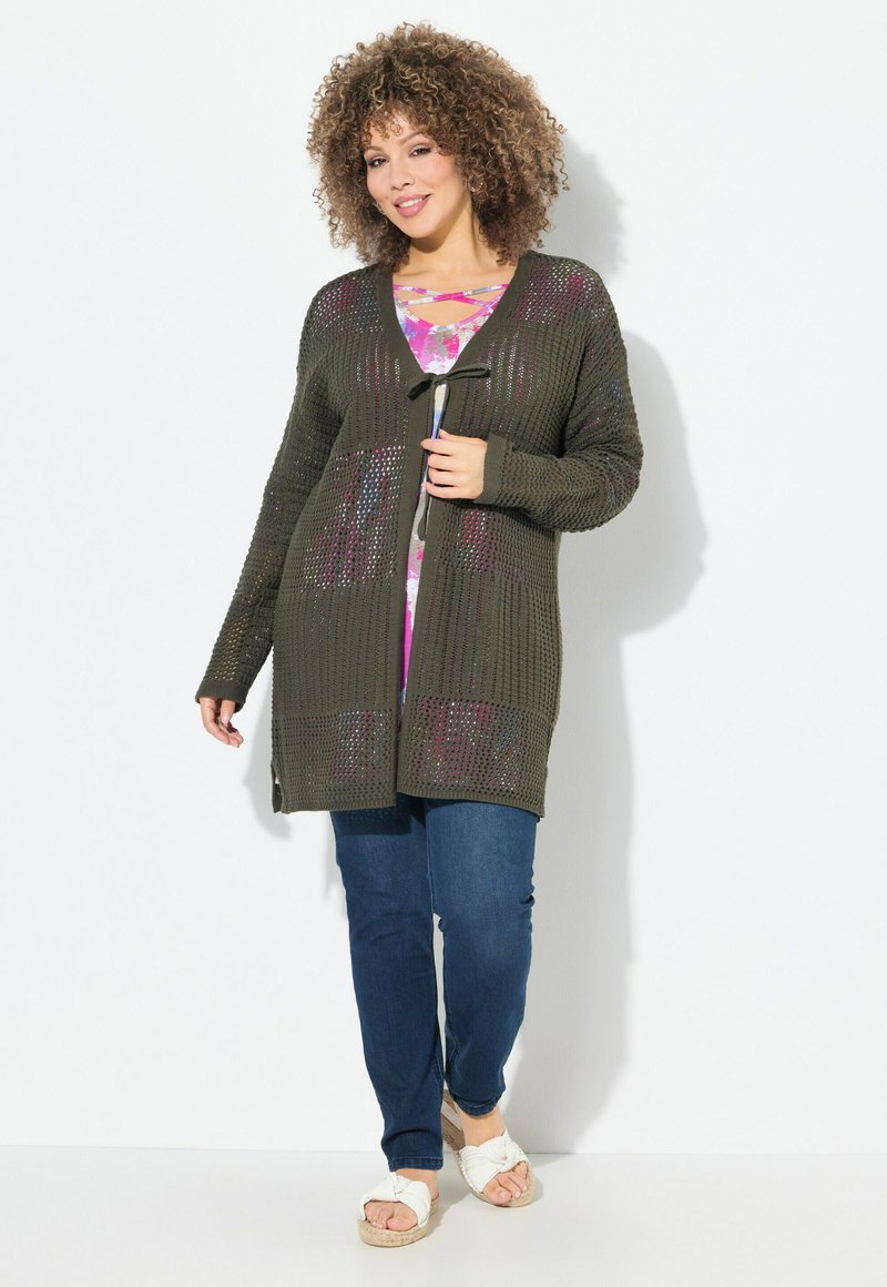 Cardigan lavorato a maglia verde oliva con trama aperta, chiusura davanti con lacci, tasche, indossato sopra una maglietta rosa a fantasia e abbinato a jeans blu e sandali.