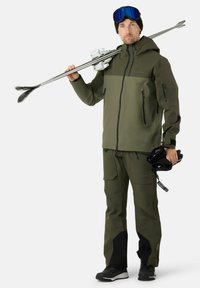 Abbigliamento da sci in verde oliva, composto da una giacca con zipper e pantaloni con numerose tasche. Tessuto scamosciato, con accenti neri e attrezzatura da sci.