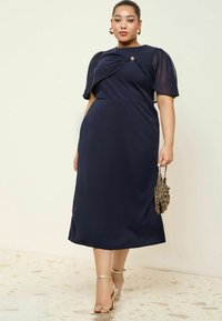 Navy Kleid mit kurzen, transparenten Puffärmeln, verdrehtem Oberteil und goldenen Akzenten. Kombiniert mit goldenen, schmalen Sandalen und einer strukturierten Tasche.