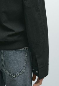 Black long-sleeve shirt with a textured surface, featuring a buttoned cuff and an angled pocket. Paired with gray denim jeans.