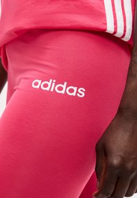 Κοντινό πλάνο ατόμου που φοράει ροζ κολάν Adidas με λευκό λογότυπο και μέρος ενός ταιριαστού τοπ με τρεις λευκές ρίγες στο πλάι.
