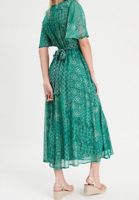 Longue robe maxi verte avec motifs floraux et géométriques, manches courtes, taille nouée, et tissu fluide. Associée à des sandales compensées marron clair.