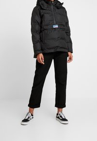 Sort puffet jakke med høj krave, lynlåslukning fortil og forlommer. Den har et logo mærke og er parret med sorte bukser og sneakers.