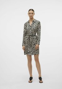 Vestito avvolgente nero e beige a fantasia con maniche lunghe, vita arricciata e colletto, abbinato a sandali neri.