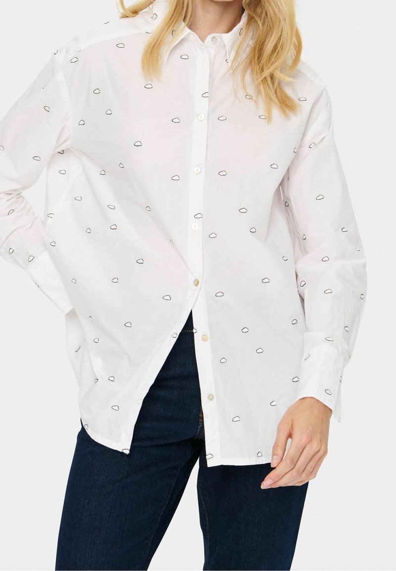 Chemise blanche à boutons avec de petits motifs de nuages noirs portée avec un jean bleu foncé, vue du cou jusqu'aux hanches.