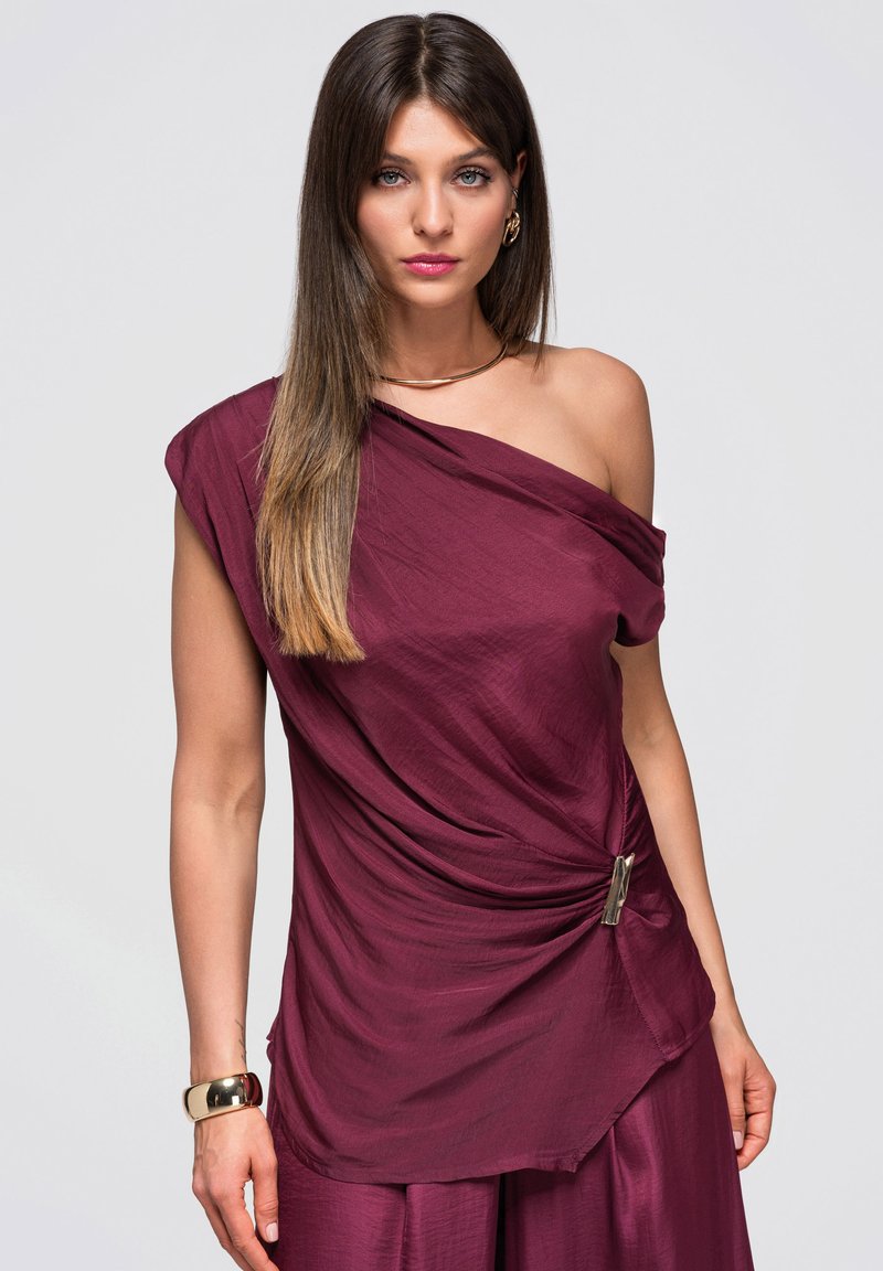 Femme aux longs cheveux bruns portant une robe bordeaux asymétrique avec une boucle métallique et des bijoux en or, debout devant un fond clair uni.