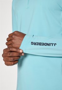 Jasnoniebieska koszula z długim rękawem i kołnierzykiem, z czarnym logo "JLINGZBERG" na mankiecie, wykonana z gładkiego materiału i dobrze dopasowana.