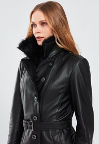 Il cappotto in pelle nera presenta un alto colletto in finta pelliccia nera, chiusura con bottoni e una vita regolabile con cintura. Texture liscia e design su misura.