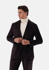 Brauner Cordblazer mit spitzem Revers und zwei vorderen Taschen, getragen über einem cremefarbenen Rollkragenpullover. Gestreifte Textur und taillierte Passform sind sichtbar.