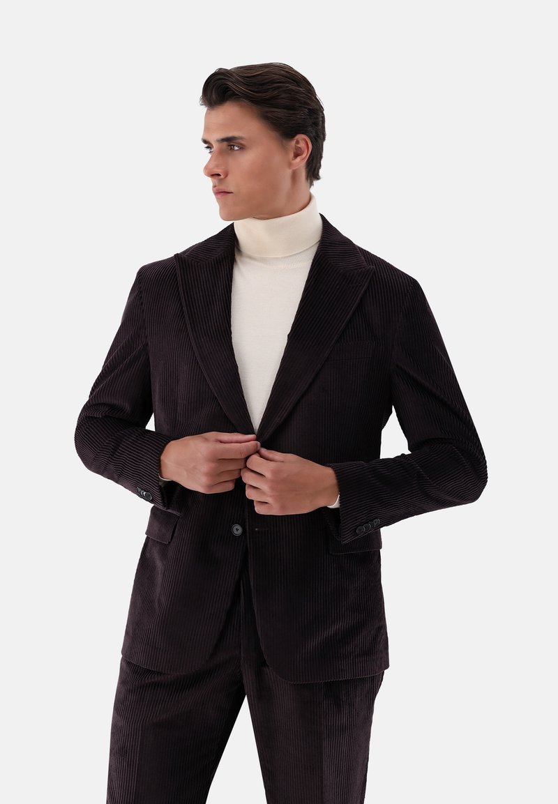 Brauner Cordblazer mit spitzem Revers und zwei vorderen Taschen, getragen über einem cremefarbenen Rollkragenpullover. Gestreifte Textur und taillierte Passform sind sichtbar.