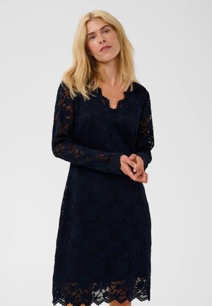 Femme blonde portant une robe en dentelle bleu marine à manches longues avec des bords festonnés, debout, les mains doucement jointes devant elle.