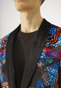 Fargerik blazer med sommerfugl-mønstre i blå, oransje og rød, med en svart satengkrage, satt sammen med en mørk, ensfarget t-skjorte.
