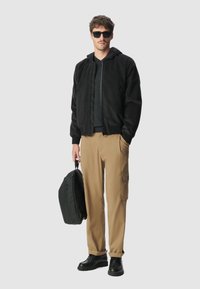 Man in een zwarte hoodie, zwarte zonnenbril, grijs shirt, beige cargobroek, zwarte schoenen, met een zwarte rugzak tegen een effen achtergrond.