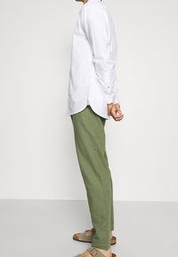 Mand iført grønne bukser, hvid langærmet skjorte og beige sandaler, der står sidelæns med hænderne bag ryggen.