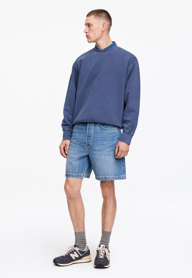 Jeune homme portant un sweat bleu, un short en jean, des chaussettes grises et des baskets bleu marine, debout et regardant sur le côté sur fond blanc.