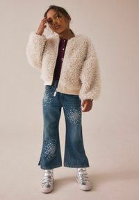 Barnet bär en krämfärgad fluffig jacka, vinröd skjorta, blå jeans med vita rhinestones-mönster och silvriga sneakers, stående mot en beige bakgrund.