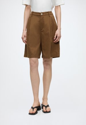 Person iført brune knælange, brede shorts, hvid kortærmet top og sorte hælede tåsandaler, stående på en hvid baggrund.