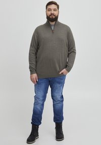Suéter verde oscuro con media cremallera, mangas largas y dobladillo acanalado, combinado con jeans azules y botas negras con cordones.
