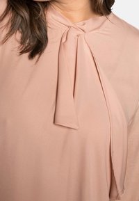 Persikofärgad blus i mjukt tyg, med knytning vid halsen och draperade ärmar, med slät textur och minimalistisk design.