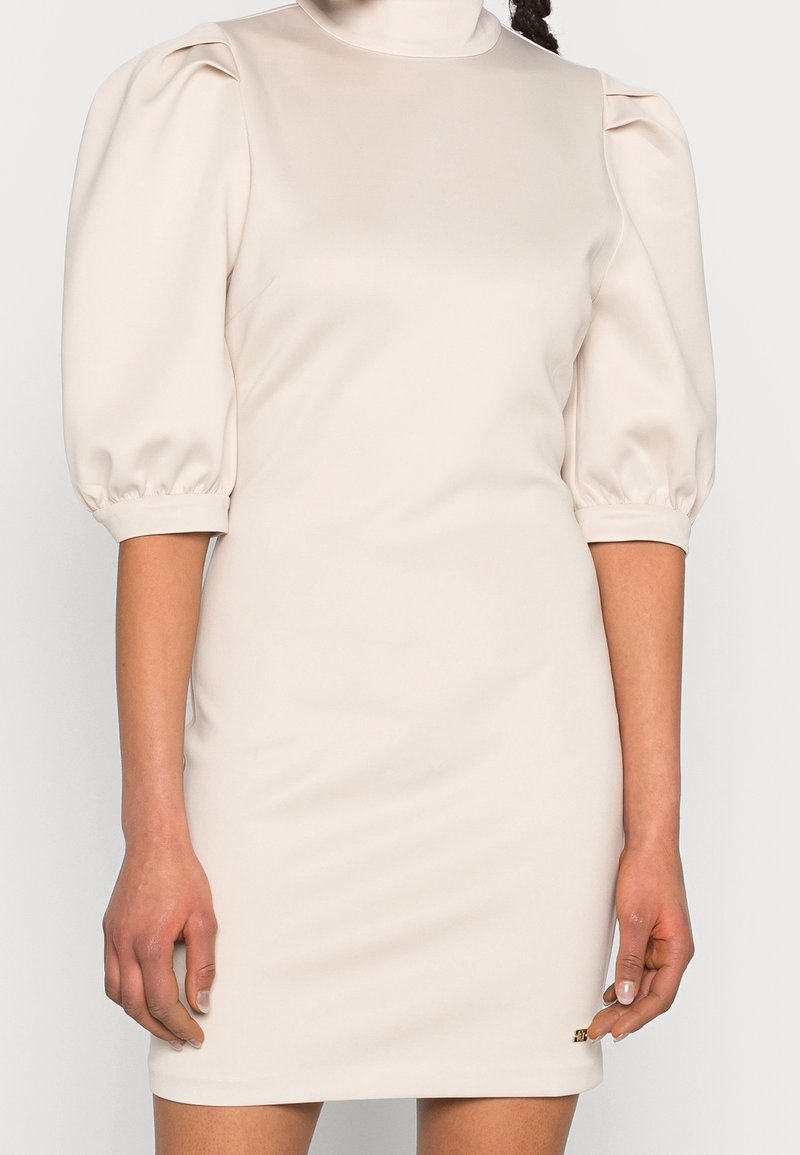 Femme portant une robe crème avec des manches bouffantes aux coudes et un col montant, mains détendues le long du corps.