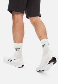 Biele športové topánky s čiernymi akcentmi, texturovaným sieťovaným zvrškom a hrubou podrážkou. Nosí sa s krémovými ponožkami s čiernym brandingom.