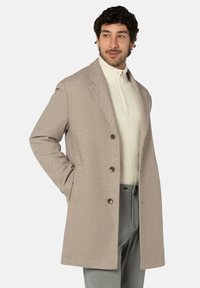 Beiger Mantel aus Wollmischung mit tailliertem Schnitt, versehen mit Kent-Kragen, drei Knöpfen und zwei Seitentaschen. Über einem strukturierten weißen Pullover getragen.