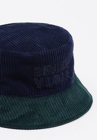Kapelusz typu bucket wykonany z korduroju z ciemnoniebieską górą i zielonym daszkiem, z wytłoczonym na przodzie logo marki. Teksturowana powierzchnia z prążkowanym wzorem.