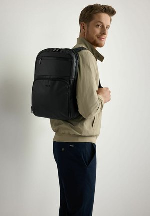 Mężczyzna w beżowej kurtce i granatowych spodniach niesie czarny plecak, patrząc przez ramię z lekkim uśmiechem na tle jednolitego tła.