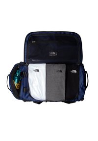Borsa a duffel blu scuro con una tasca in rete. Contiene camicie piegate di colore bianco, grigio e nero. Include una borraccia blu con logo.