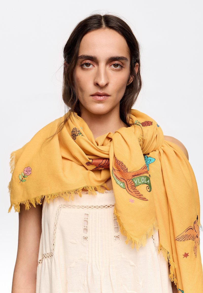 Yellow scarf with embroidered birds and flowers; soft texture, fringed edges, and colorful detailing. Worn loosely around the neck.
