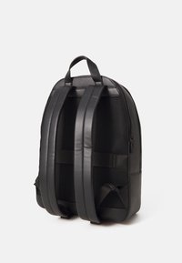 Sac à dos noir avec un extérieur en cuir lisse et des panneaux latéraux en mesh. Équipé de larges sangles rembourrées et d'une poignée supérieure pour le transport.