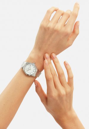 Montre analogique argentée avec un cadran blanc rond, un bracelet en métal et des index horaires numériques, portée sur un poignet à la peau de couleur neutre.