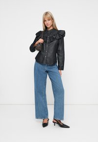 Svart denimskjorte med volanger på skuldrene, knapp-up design, kombinert med vide blå jeans og svarte spisse hæler, holder en liten veske.