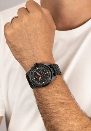 Musta metallist käekell ümmarguse näo, indeksi markerite, punaste aktsentidega ja lingitud rihmaga. Kandmisel heledate pruunide juustega randmel.