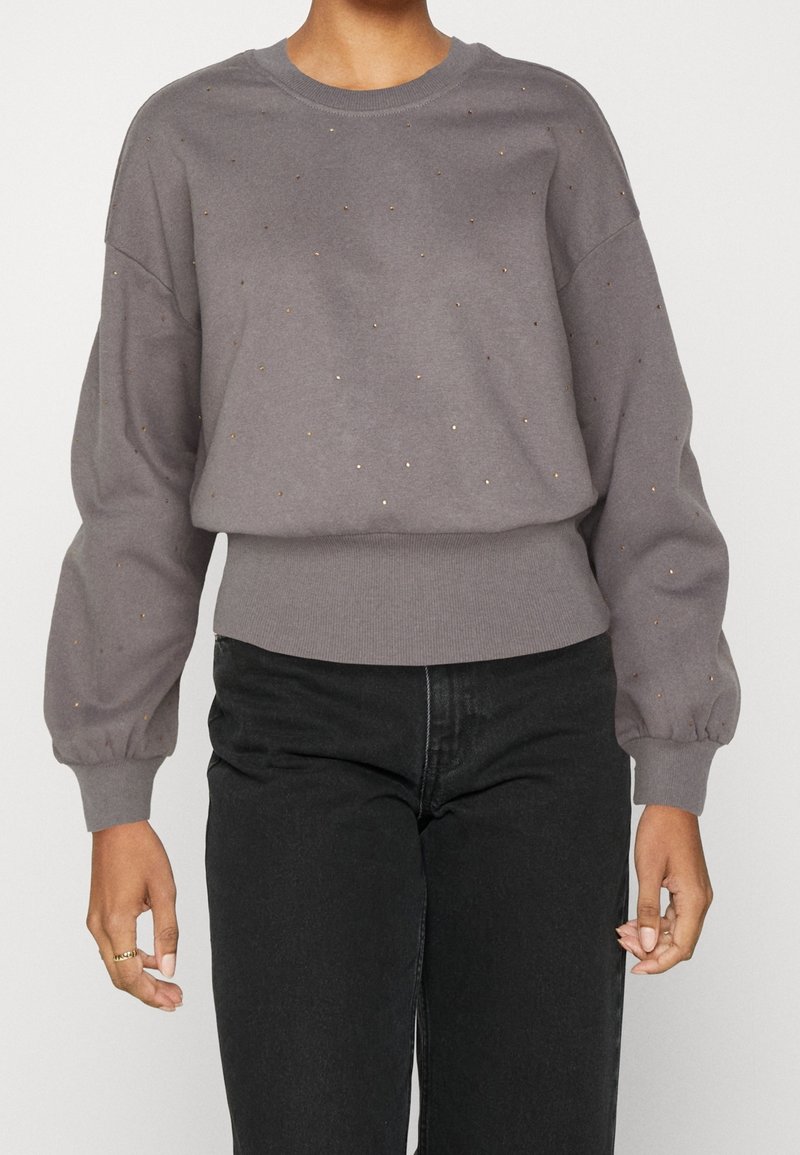 Personne portant un pull gris à manches longues avec de petits clous métalliques et un jean noir taille haute, debout les bras détendus.