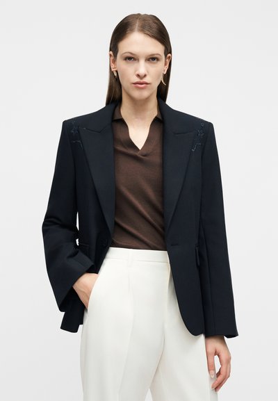 Γυναίκα που φοράει μαύρο σακάκι με κεντημένα αστέρια, καφέ τοπ με v-λαιμόκοψη και λευκό παντελόνι ψηλόμεσο, στέκεται με το ένα χέρι στην τσέπη.