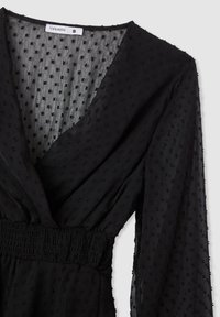 Vestito nero con scollatura a V, realizzato in tessuto testurizzato con piccole pois e vita arricciata. Maniche lunghe e fluide arricchiscono il design.